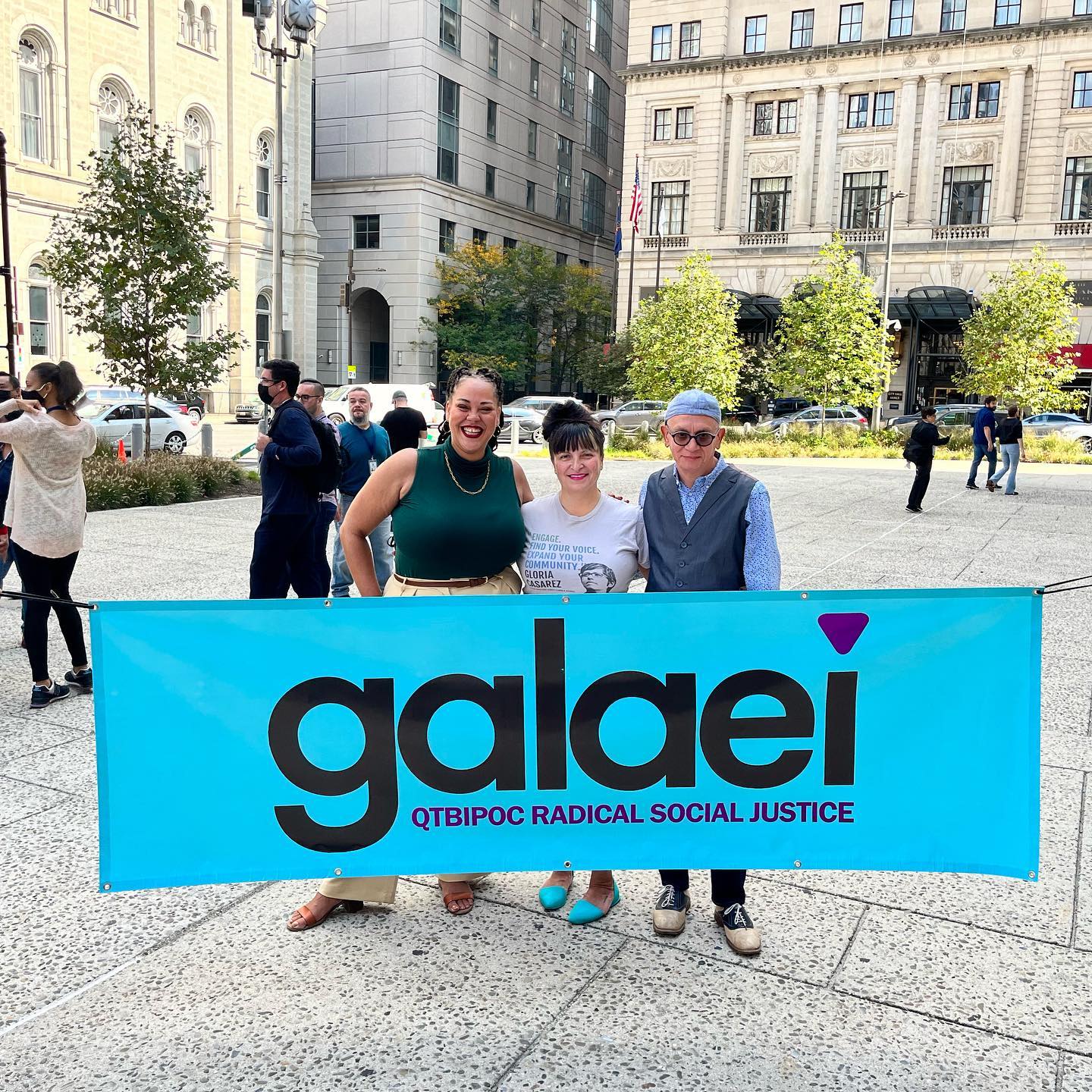 Three GALAEI volunteers in Philadelphia in front of a light blue banner that says: GALAEI: QTBIPOC Radical Social Justice